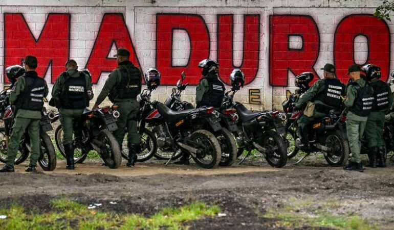 Members of the National Guard stand guard in front of a mural of ousted Venezuela's President Nicolas Maduro in Caracas on January 10, 2026. The US State Department on January 10, urged Americans in Venezuela to leave the country "immediately," citing risks from armed militias searching vehicles for US citizens at roadblocks. (Photo by Juan BARRETO / AFP via Getty Images)