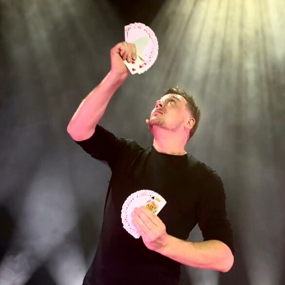 Magician Jamie Allan holding fanned playing cards, looking up at cards in his raised hand.
