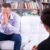 A man in distress sits on a couch during a therapy session, with a therapist across from him.
