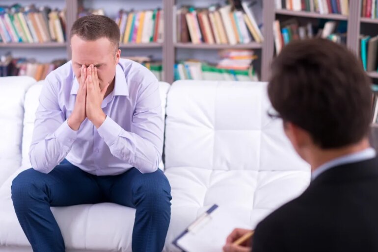 A man in distress sits on a couch during a therapy session, with a therapist across from him.