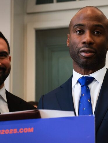 Zohran Mamdani, mayor-elect, smiles next to a man wearing a blue tie with colorful patterns, standing behind a podium that says "Transition2025.com".