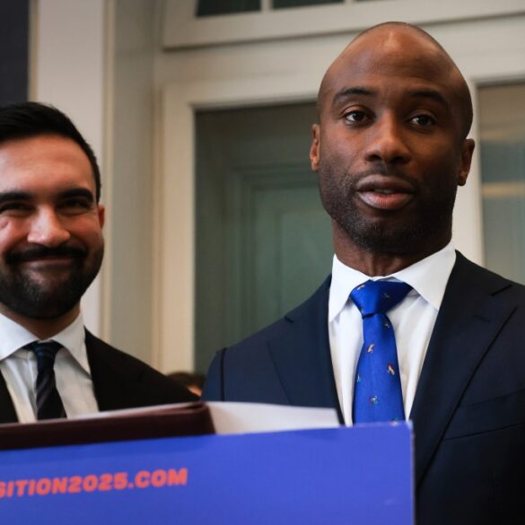 Zohran Mamdani, mayor-elect, smiles next to a man wearing a blue tie with colorful patterns, standing behind a podium that says "Transition2025.com".