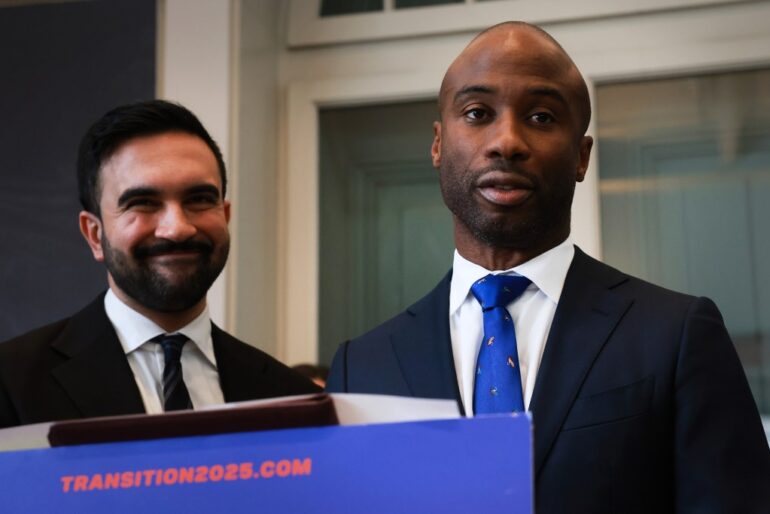 Zohran Mamdani, mayor-elect, smiles next to a man wearing a blue tie with colorful patterns, standing behind a podium that says "Transition2025.com".