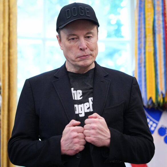 Elon Musk in a black "DOGE" baseball cap and a black t-shirt with "the dogef" printed on it, in the White House's Oval Office.