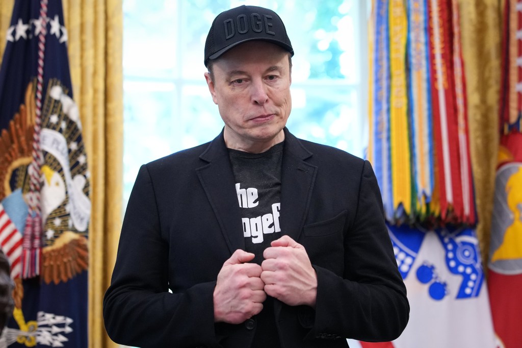 Elon Musk in a black "DOGE" baseball cap and a black t-shirt with "the dogef" printed on it, in the White House's Oval Office.
