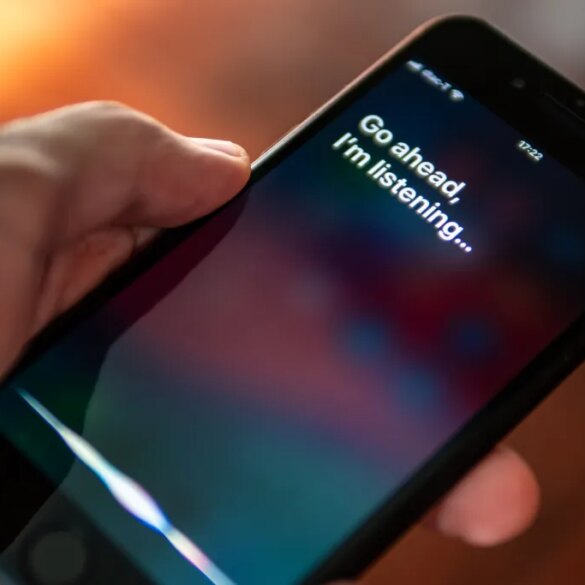 A hand holding an iPhone with the Siri interface showing the text "Go ahead, I'm listening..."