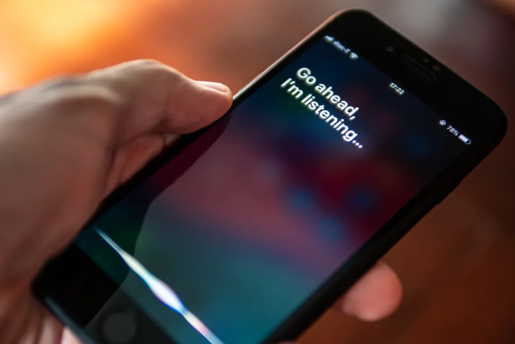 A hand holding an iPhone with the Siri interface showing the text "Go ahead, I'm listening..."
