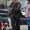 Will Ferrell in costume as a character with long blonde hair and a beard, wearing a black sheer shirt, a red and black vest, and black leather pants, on a film set.