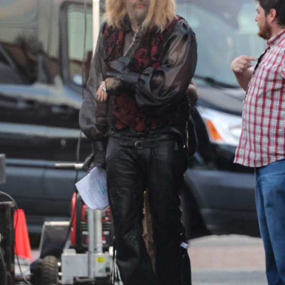 Will Ferrell in costume as a character with long blonde hair and a beard, wearing a black sheer shirt, a red and black vest, and black leather pants, on a film set.