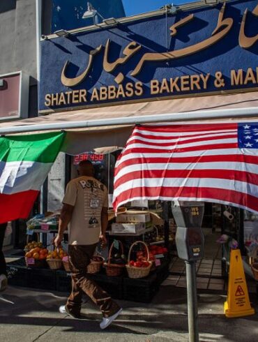 A man walks by a Iranian Market with an Iranian and an American flag outsite the shop in the Persian Square at the West LA neighborhood with in Los Angeles, California on February 28, 2026. The US and Israel launched an attack of unprecedented scale against Iran on Saturday, reportedly killing more than 200 people, with Tehran launching a retaliatory missile barrage that sent people running for cover across the Middle East. (Photo by Apu GOMES / AFP via Getty Images)