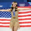 Gold medalist Alysa Liu of Team United States, in a sparkling gold dress, holds an American flag behind her shoulders during the medal ceremony.