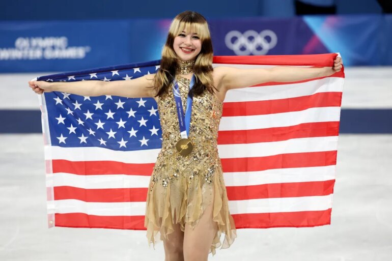 Gold medalist Alysa Liu of Team United States, in a sparkling gold dress, holds an American flag behind her shoulders during the medal ceremony.