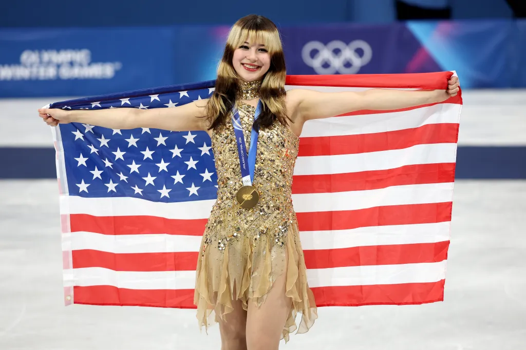 Gold medalist Alysa Liu of Team United States, in a sparkling gold dress, holds an American flag behind her shoulders during the medal ceremony.
