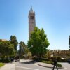 A young male student walks past Sather Tower and other campus buildings on a sunny day at UC Berkeley.
