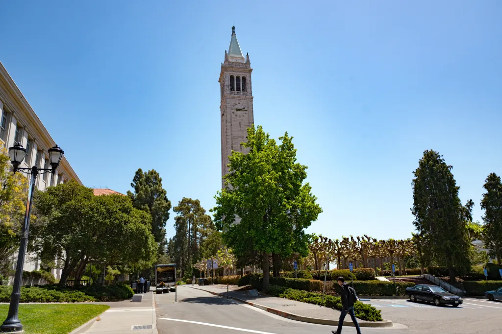 A young male student walks past Sather Tower and other campus buildings on a sunny day at UC Berkeley.