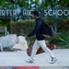A student walks past the "Charter High School" sign carrying a backpack and a violin case.