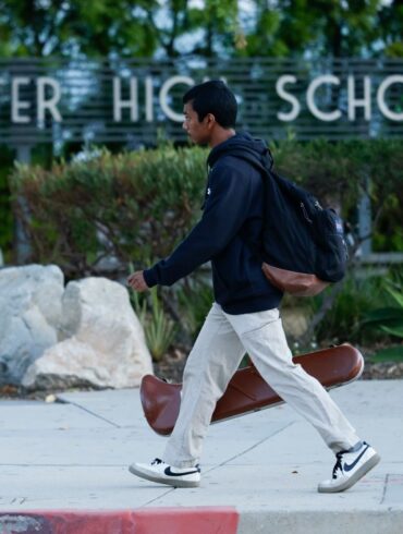 A student walks past the "Charter High School" sign carrying a backpack and a violin case.