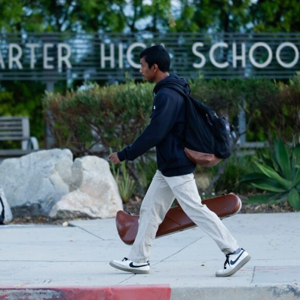 A student walks past the "Charter High School" sign carrying a backpack and a violin case.
