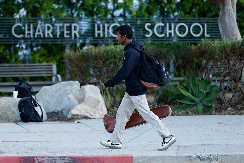 A student walks past the "Charter High School" sign carrying a backpack and a violin case.