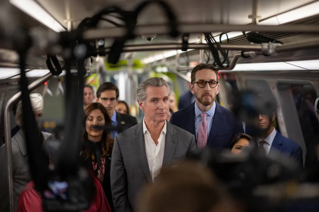 California Governor Gavin Newsom on a train, surrounded by a crowd and cameras.