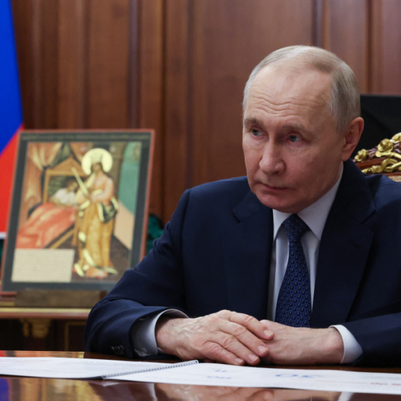 Vladimir Putin seated at a desk, looking to his left, with the Russian flag and an icon in the background.
