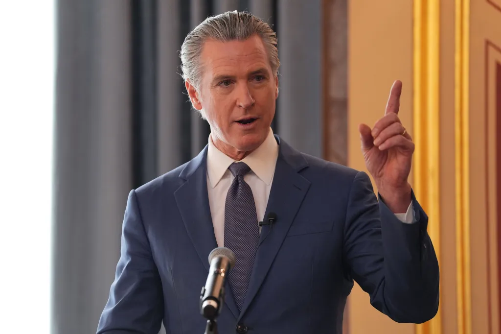 California Governor Gavin Newsom speaking into a microphone, pointing with his right index finger.