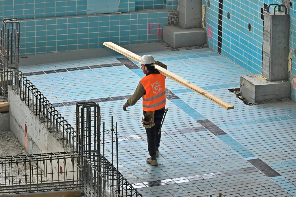 A construction worker in a hard hat and safety vest carries a wooden plank across a partially tiled swimming pool undergoing renovation.