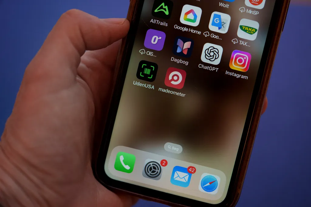 A close-up of a hand holding a smartphone displaying apps, including "UdenUSA" and "Madeometer", designed to filter out American products.