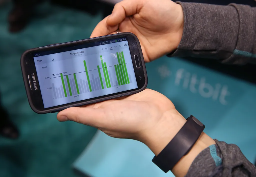 Hands holding a Samsung smartphone displaying a Fitbit Force bar graph of steps walked, with a Fitbit Force fitness tracker on one wrist.