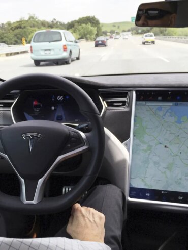 The interior of a Tesla Model S in San Francisco, showing the steering wheel and a large center screen displaying a map, with a highway visible through the windshield.