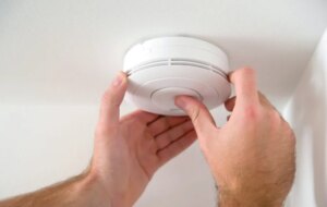 A person installing a white smoke or carbon monoxide detector on a white ceiling.