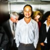 A smiling young man in an elevator as four other business people around him hold their noses.