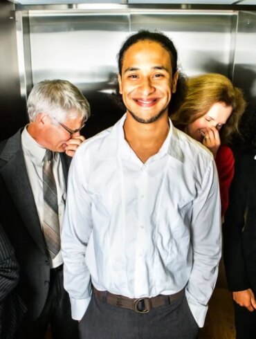 A smiling young man in an elevator as four other business people around him hold their noses.
