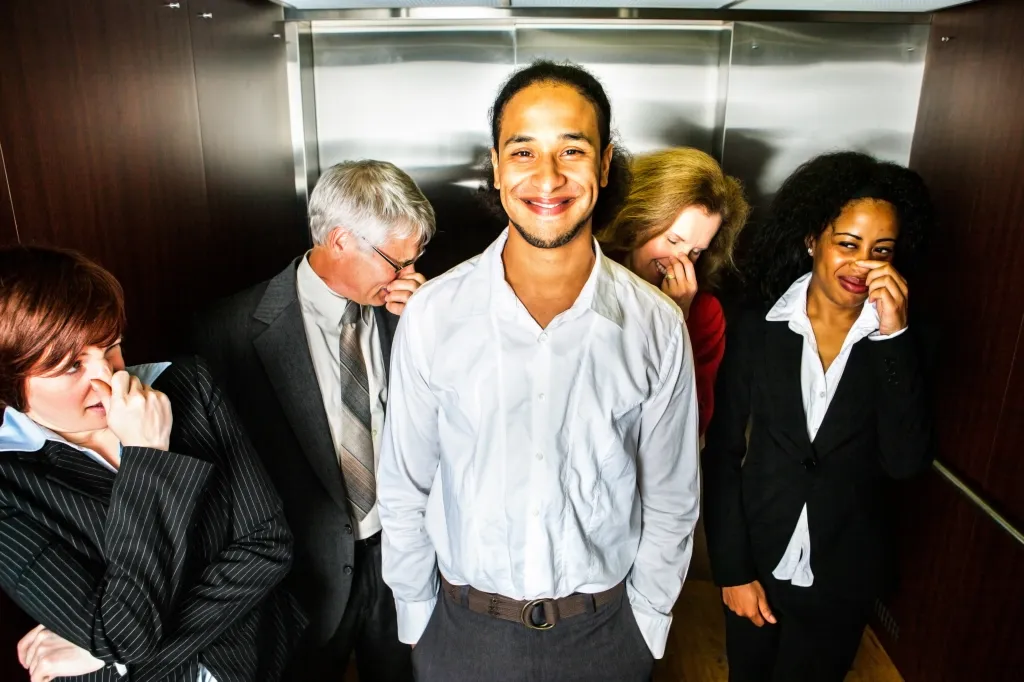 A smiling young man in an elevator as four other business people around him hold their noses.