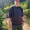 Chuck Norris wearing a black cap, gray shirt, and green shorts, standing on a hiking trail.