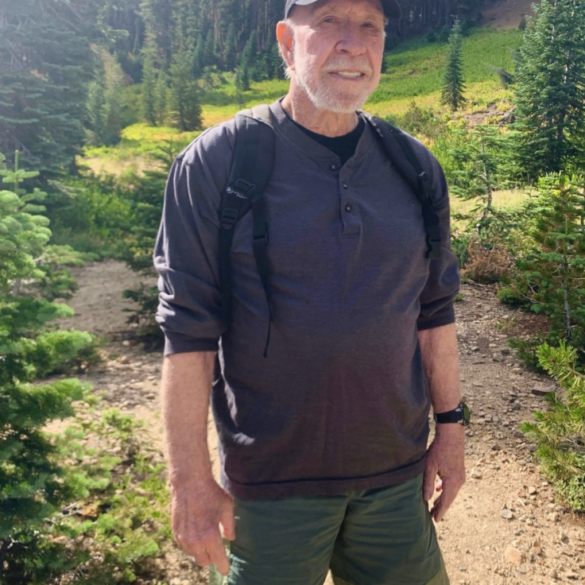 Chuck Norris wearing a black cap, gray shirt, and green shorts, standing on a hiking trail.