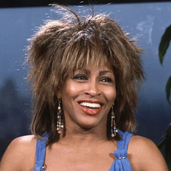 Tina Turner smiling with her mouth open, wearing a blue top and beaded earrings.
