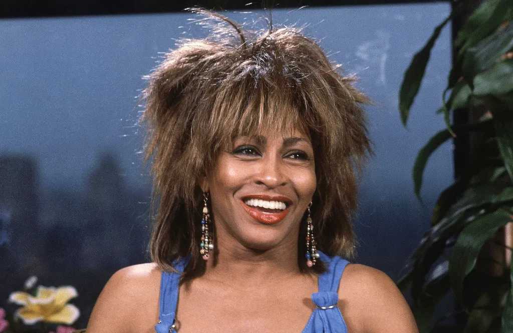 Tina Turner smiling with her mouth open, wearing a blue top and beaded earrings.