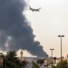 An Emirates aircraft prepares for landing as a smoke plume rises from an ongoing fire near Dubai International Airport in Dubai on March 16, 2026. Flights were gradually resuming at Dubai airport on March 16, previously the world's busiest for international flights, the airport operator said, after a "drone-related incident" sparked a fuel tank fire nearby, as Iran kept up its Gulf attacks. (Photo by AFP via Getty Images) /