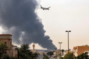 An Emirates aircraft prepares for landing as a smoke plume rises from an ongoing fire near Dubai International Airport in Dubai on March 16, 2026. Flights were gradually resuming at Dubai airport on March 16, previously the world's busiest for international flights, the airport operator said, after a "drone-related incident" sparked a fuel tank fire nearby, as Iran kept up its Gulf attacks. (Photo by AFP via Getty Images) /
