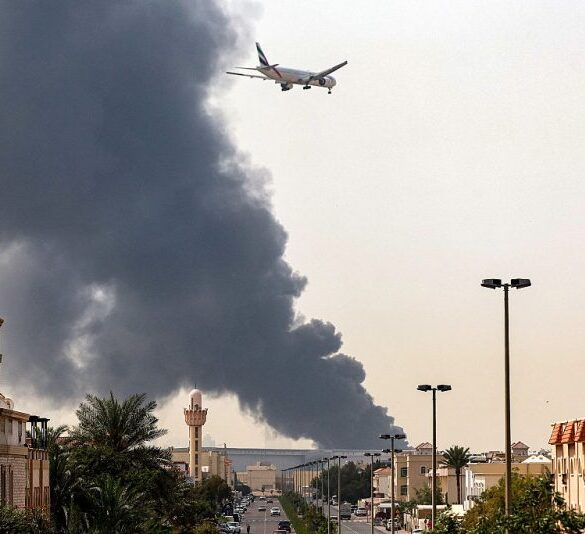 An Emirates aircraft prepares for landing as a smoke plume rises from an ongoing fire near Dubai International Airport in Dubai on March 16, 2026. Flights were gradually resuming at Dubai airport on March 16, previously the world's busiest for international flights, the airport operator said, after a "drone-related incident" sparked a fuel tank fire nearby, as Iran kept up its Gulf attacks. (Photo by AFP via Getty Images) /