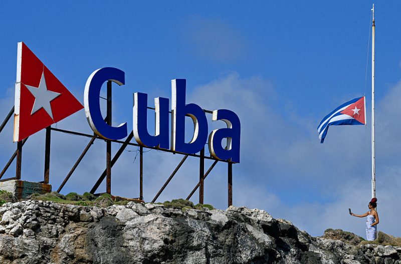 TOPSHOT - A tourist takes a selfie picture as she stands next to a Cuban national flag at half-mast in Havana on January 5, 2026. Havana declared two days of national mourning as of January 5 after a total of 32 Cubans were killed during the US attack on Caracas that culminated in the capture of Venezuela's president Nicolas Maduro. (Photo by Adalberto ROQUE / AFP via Getty Images)