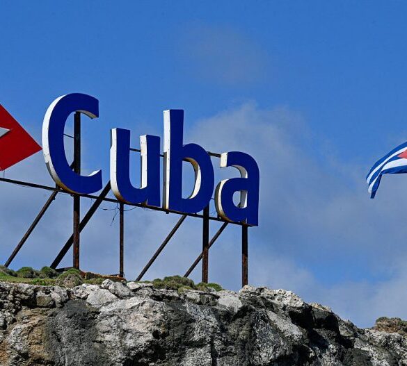 TOPSHOT - A tourist takes a selfie picture as she stands next to a Cuban national flag at half-mast in Havana on January 5, 2026. Havana declared two days of national mourning as of January 5 after a total of 32 Cubans were killed during the US attack on Caracas that culminated in the capture of Venezuela's president Nicolas Maduro. (Photo by Adalberto ROQUE / AFP via Getty Images)
