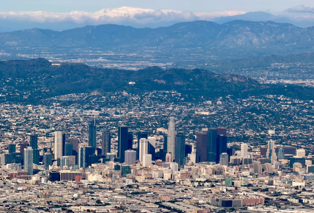 Aerial view of downtown Los Angeles with the Hollywood sign on a distant mountain and additional mountains in the background.