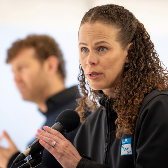 Janisse Quinones speaking at a press conference, with a man blurred in the background.