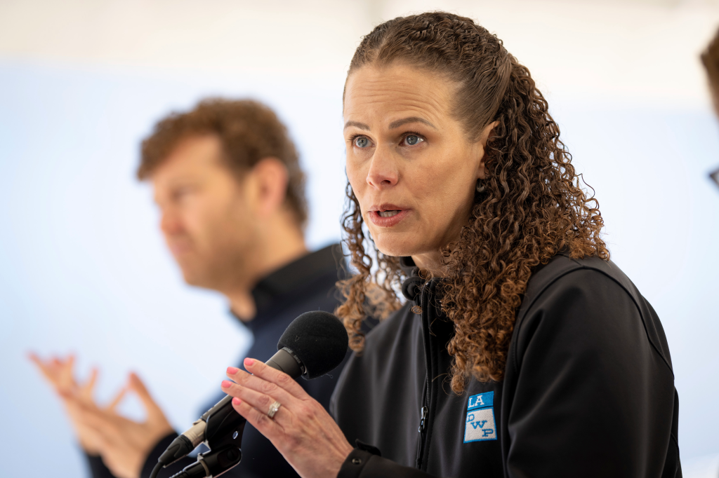 Janisse Quinones speaking at a press conference, with a man blurred in the background.