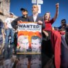 Steve Hilton, running for Governor of California, holds a "WANTED: SOME LEADERSHIP" sign depicting Karen Bass and Gavin Newsom, while a woman beside him raises her fist during a protest against wildfire response in Pacific Palisades.