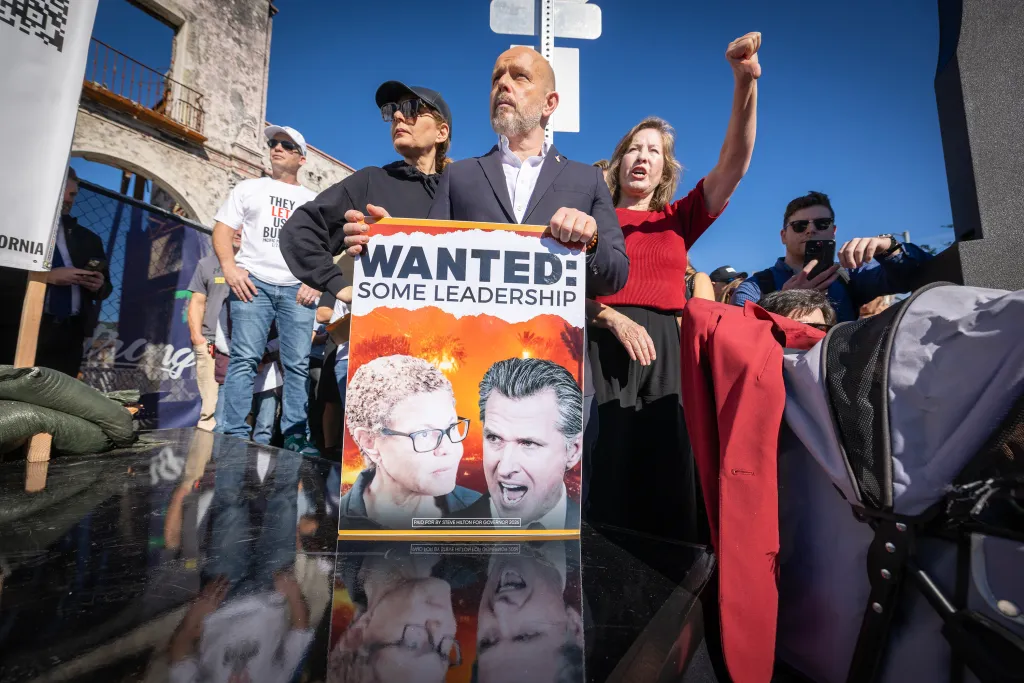 Steve Hilton, running for Governor of California, holds a "WANTED: SOME LEADERSHIP" sign depicting Karen Bass and Gavin Newsom, while a woman beside him raises her fist during a protest against wildfire response in Pacific Palisades.