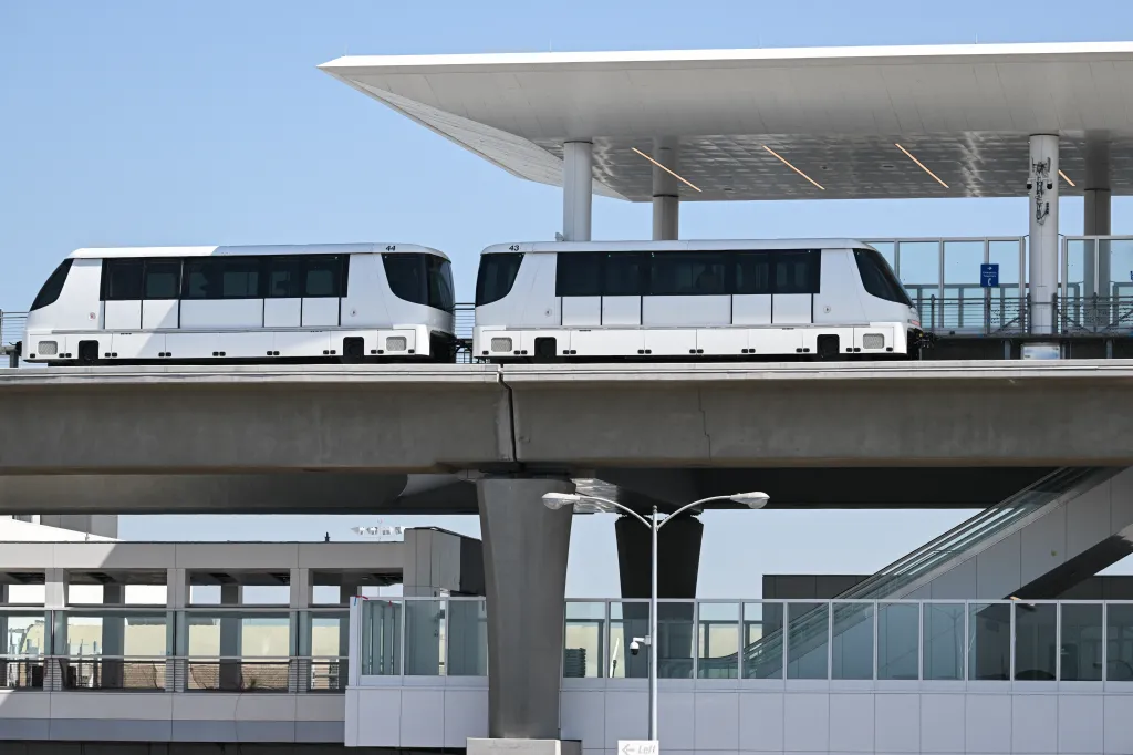Two white Automated People Mover (APM) vehicles on an elevated track approaching a station.