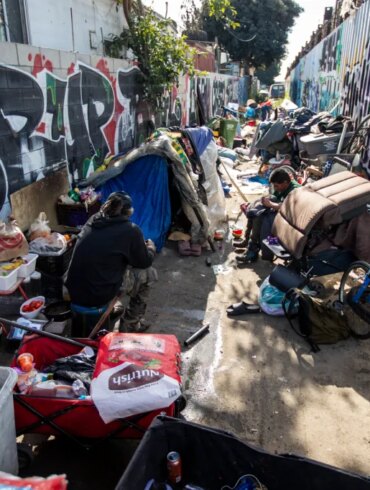 People sit among their belongings at an encampment in an alley.